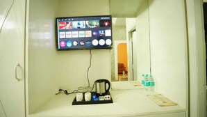Habitación de hotel con televisor de pared mostrando varios canales, cafetera, bandeja con tazas y tetera, además de una botella de agua sobre la encimera.