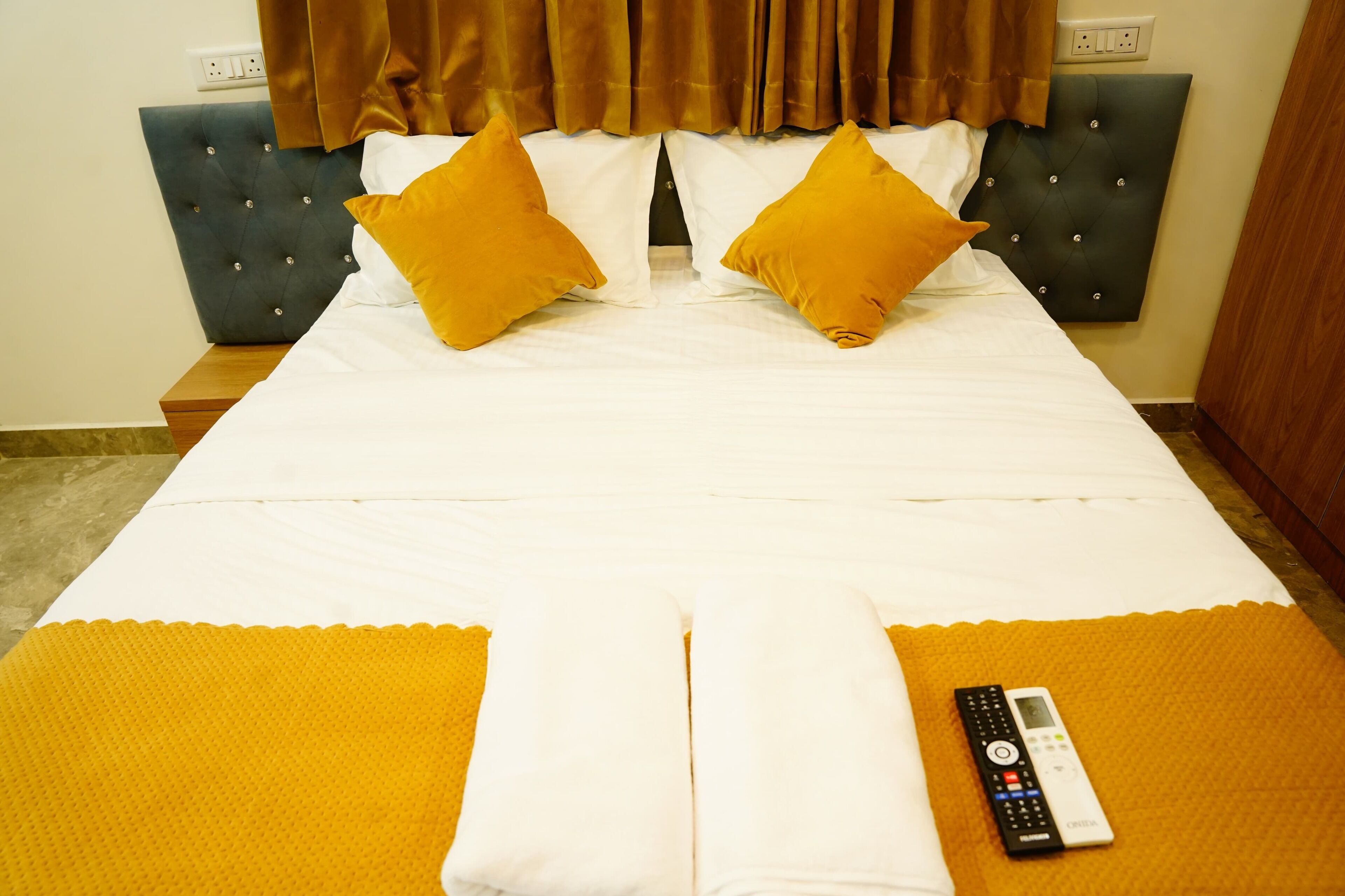 Uma cama de casal com roupa de cama branca e dois travesseiros mostarda, um controle remoto sobre um tapete amarelo, e uma cabeceira escura.