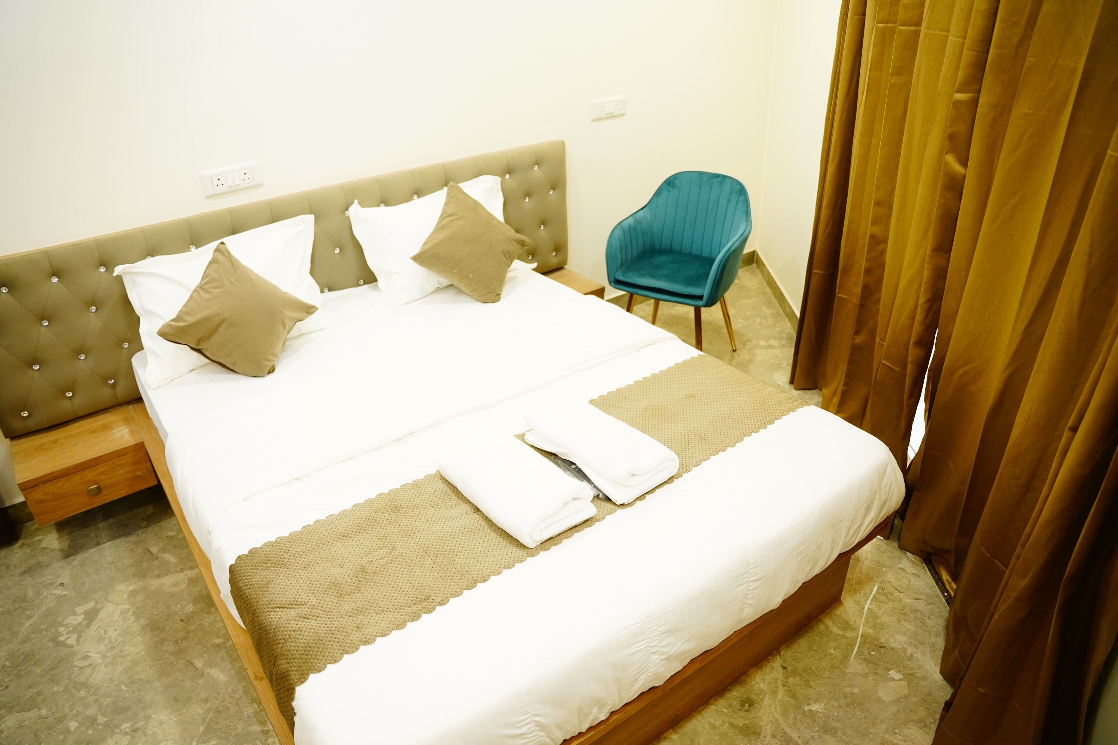A neatly made bed with a tufted headboard, a blue chair, and a brown curtain.