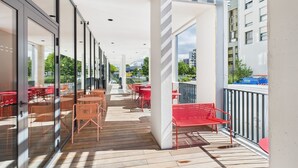 Un espace extérieur abrité, aménagé avec des tables et des chaises, une terrasse en bois et offrant une vue sur les bâtiments et les arbres.