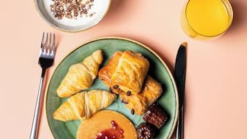 Een ontbijttafel met croissants, pannenkoeken, jam, een kop koffie, een kom granola en een glas sinaasappelsap.
