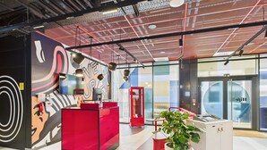 Un espace d’accueil moderne avec un bureau rouge, une grande fresque murale et une plante en pot.
