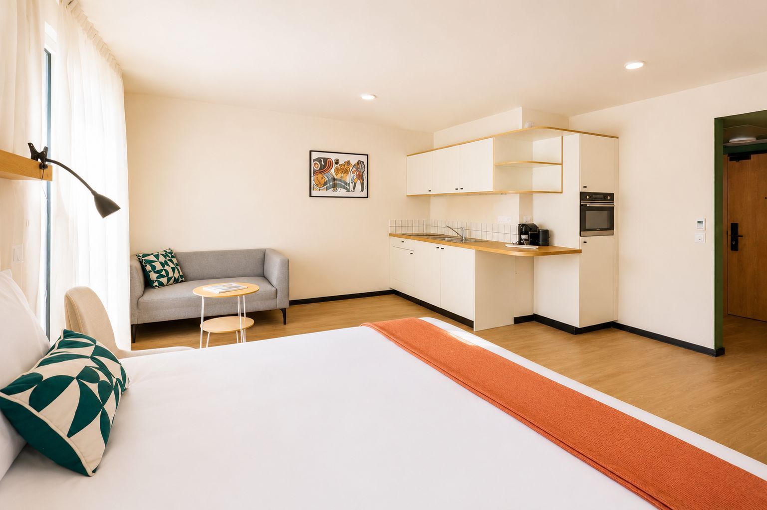 A hotel room with a bed, a sofa, a small table, a kitchenette, and a painting on the wall.
