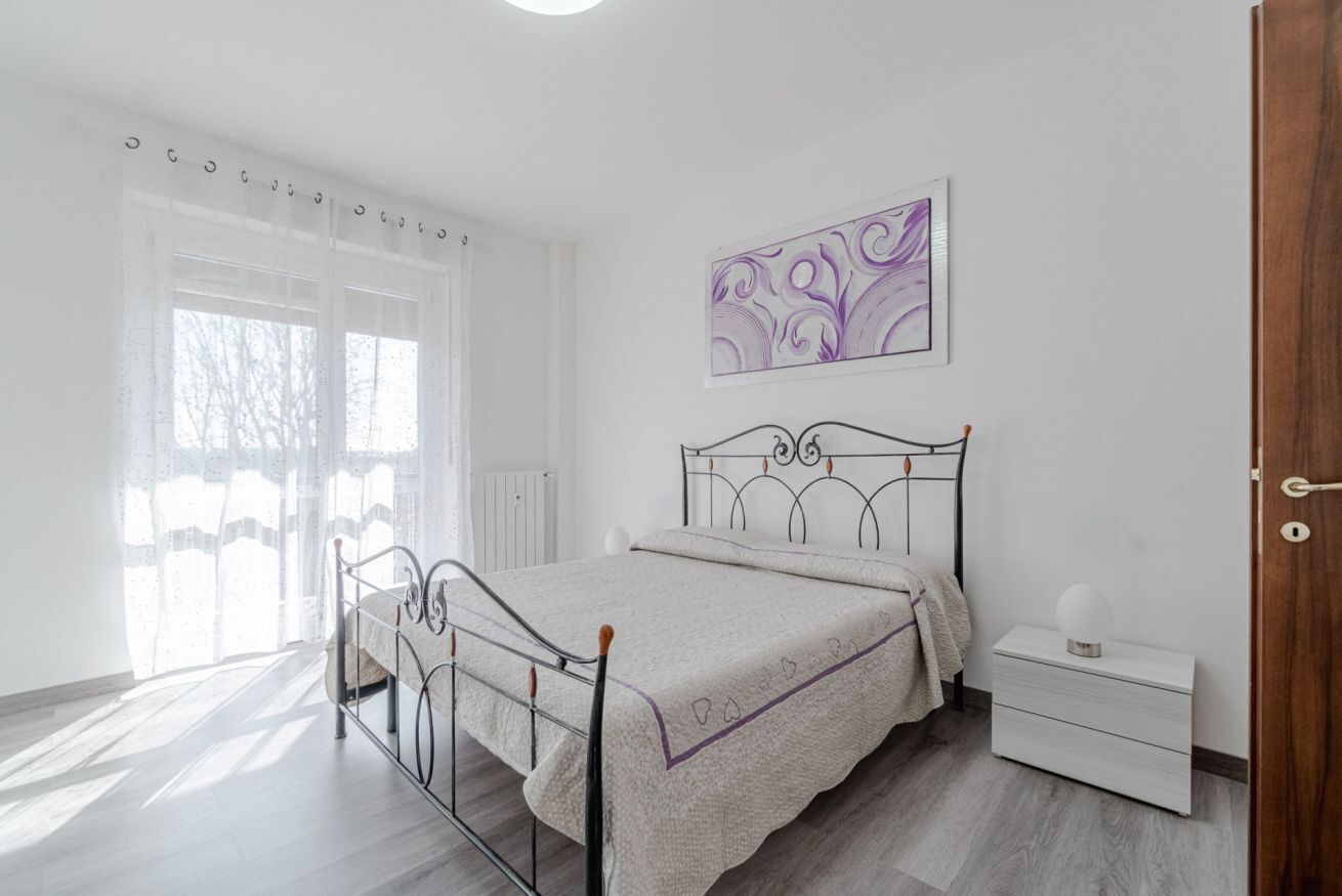 A bedroom with a metal bed frame, a bedside table, a nightstand, a window with sheer curtains, and a wall-mounted artwork.