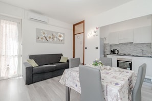 Una sala de estar moderna con un sofá gris, una mesa de comedor con mantel floral y una cocina con armarios blancos y salpicadero de ladrillo.