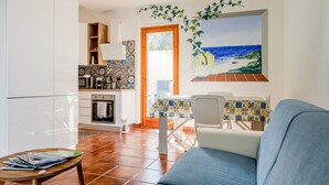 Un soggiorno con divano, un tavolo da pranzo con sedie, angolo cottura con mobili e fornelli, e un quadro raffigurante una spiaggia.