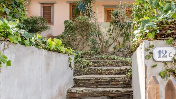 Una scala in pietra conduce a un edificio con una targa con il numero '12', immerso nel verde e circondato da un muro bianco.