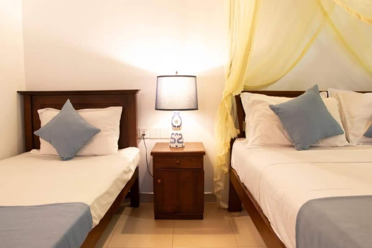 A double room with two beds, each with a wooden headboard, a bedside table with a lamp, and a yellow canopy over one bed.