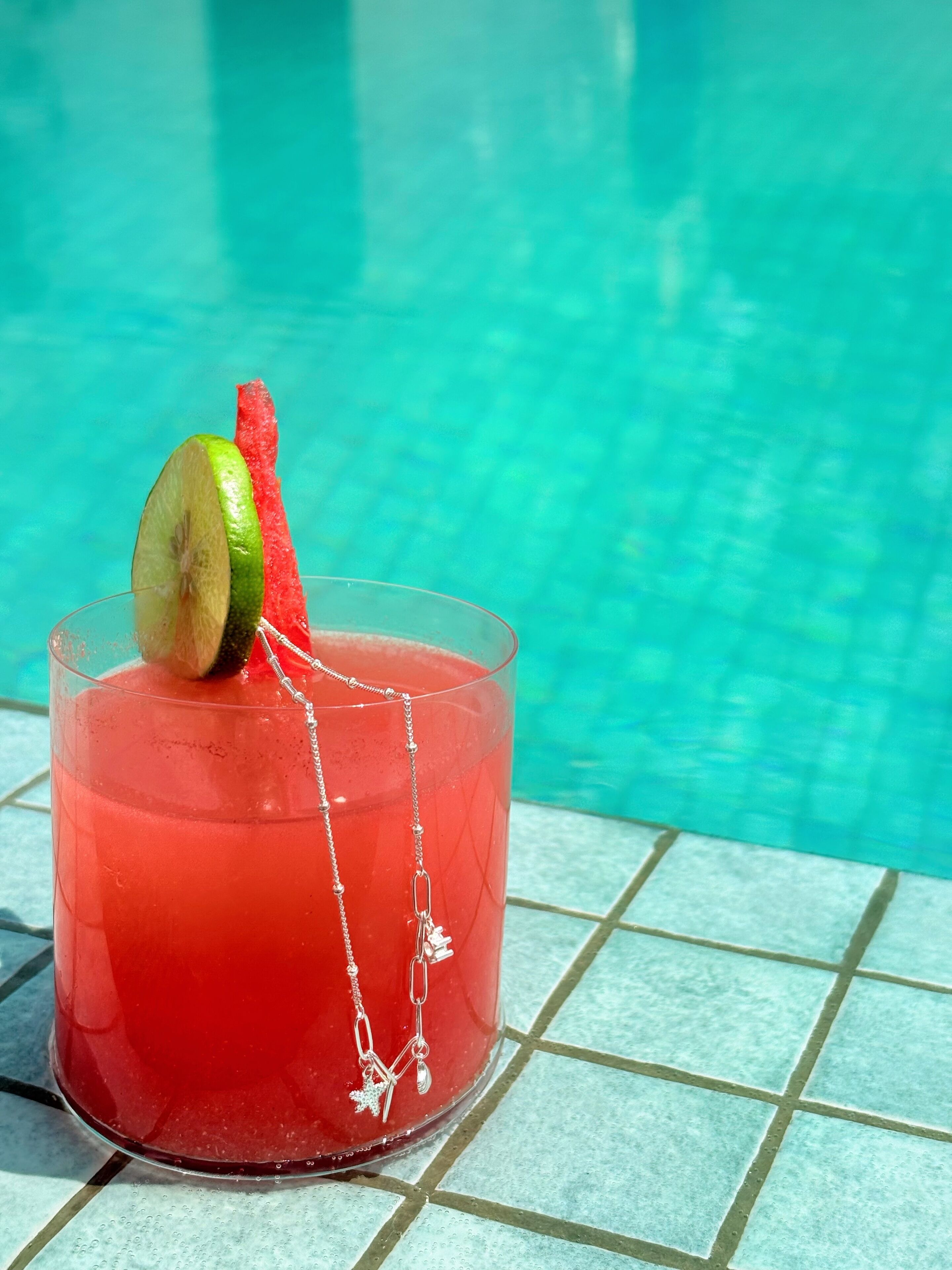 Coquetel com rodela de limão e pedaços de melancia, em um ambiente de piscina com piso de azulejos.