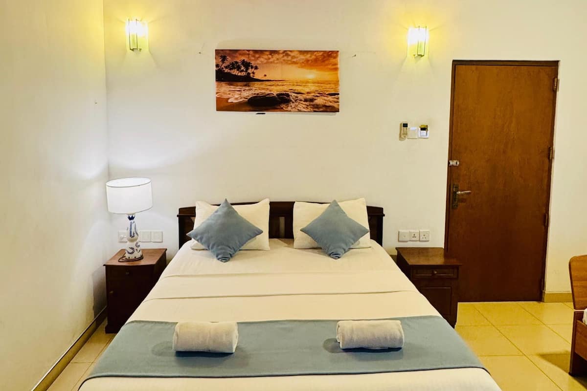 A bedroom with a wooden ceiling, a bed with two pillows, a nightstand with a lamp, a wall-mounted picture, and a door.