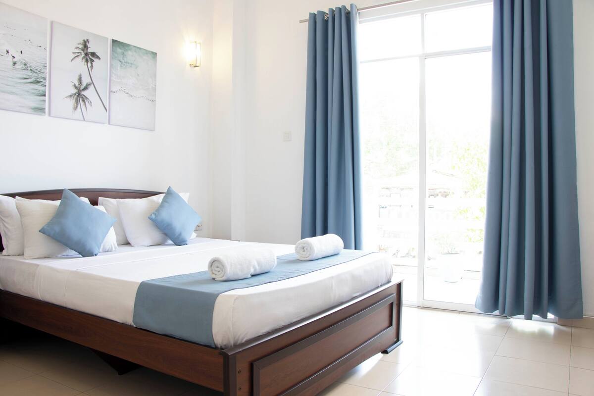 A bedroom with a large bed, a view of the outdoors through a sliding glass door, and a wall with three framed pictures.
