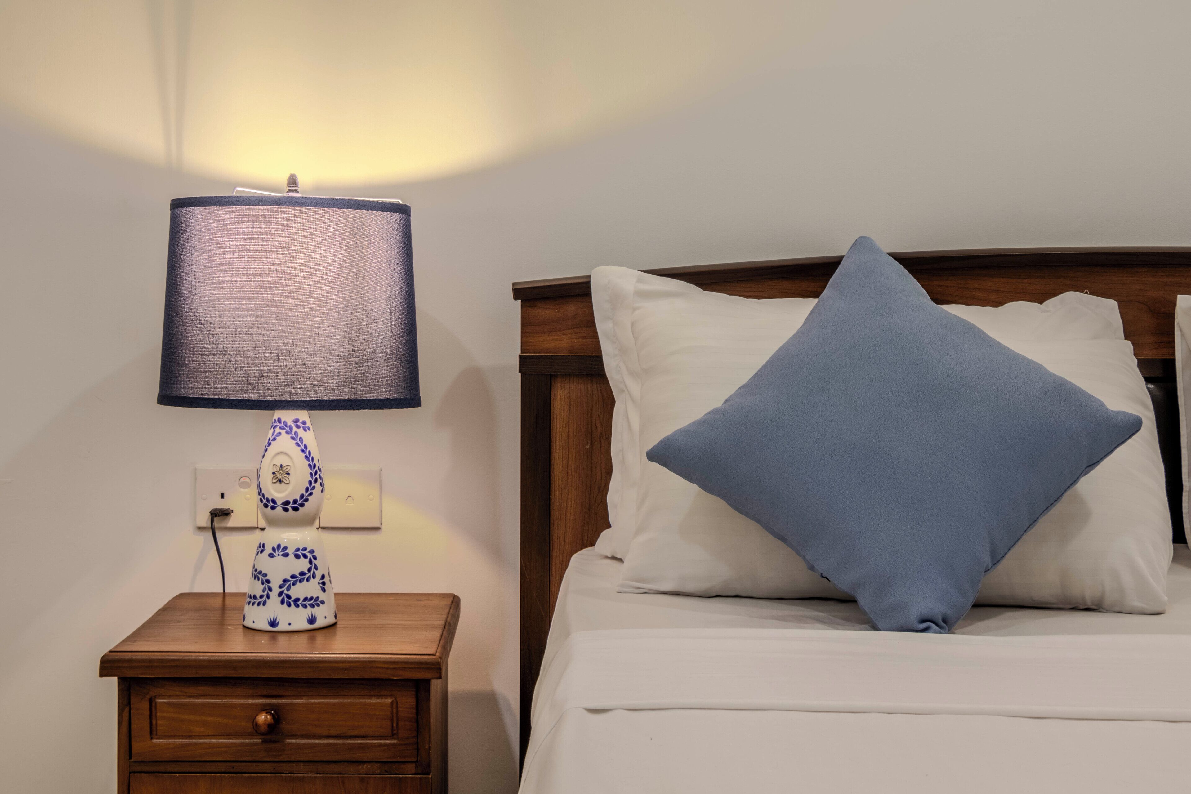 A neatly made bed with a blue pillow and a white pillow, a bedside table with a lamp, and a wooden headboard.