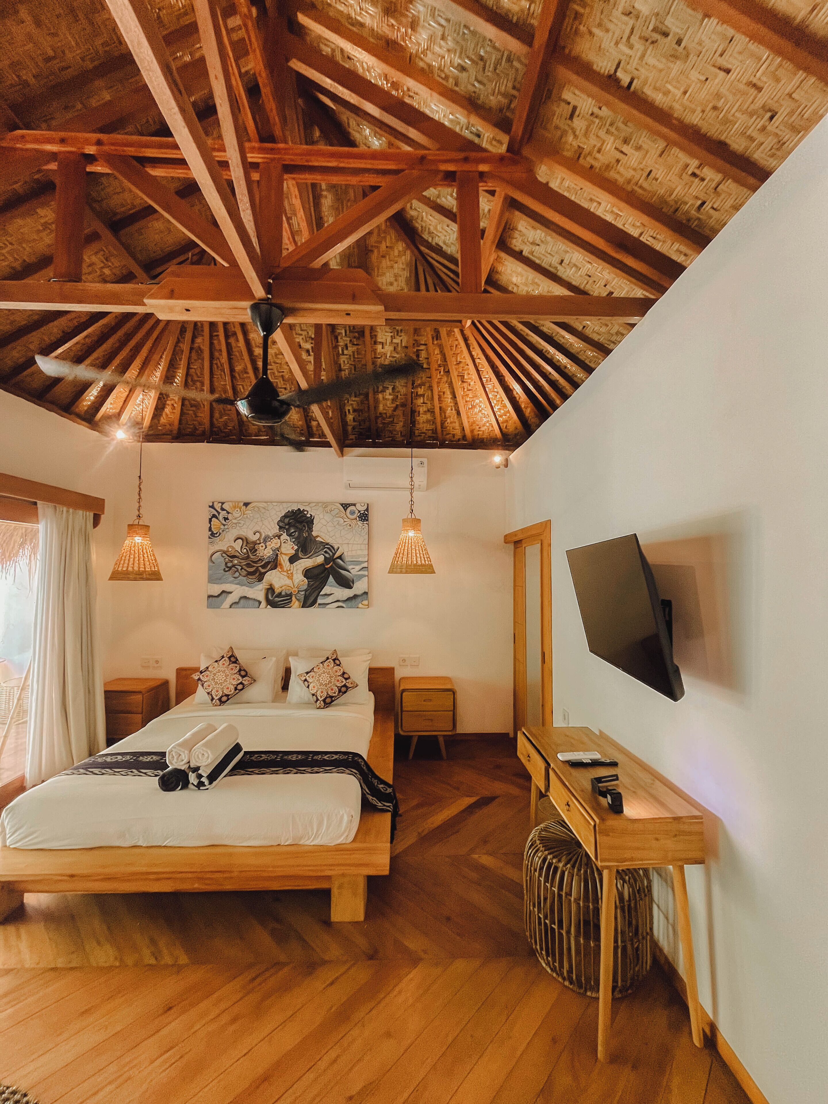 A bedroom with a wooden ceiling, a bed with pillows, a bedside table, a TV, and a hanging light fixture.