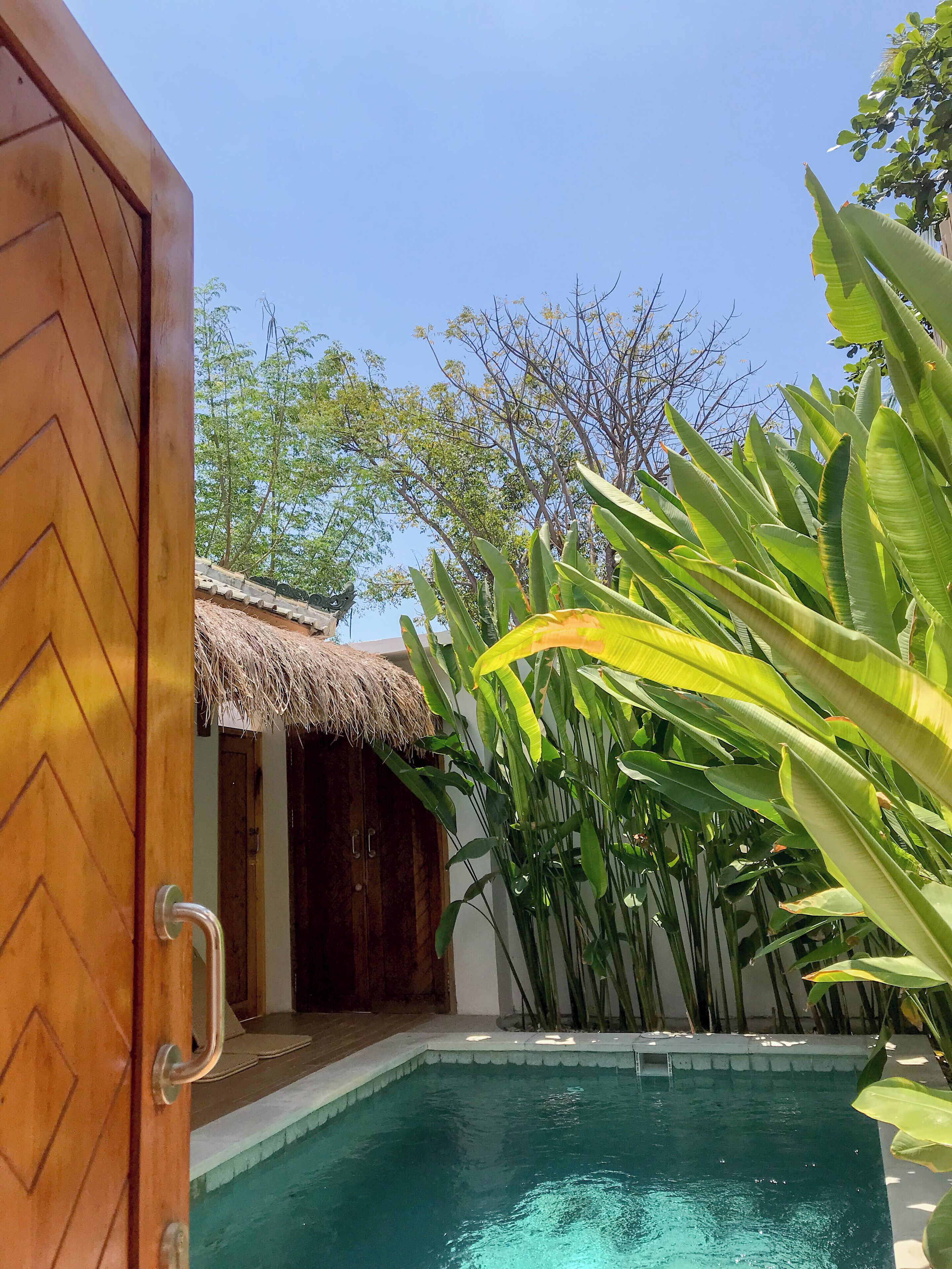 Una porta de fusta que dona a una zona de piscina amb una construcció de teules i vegetació exuberant.