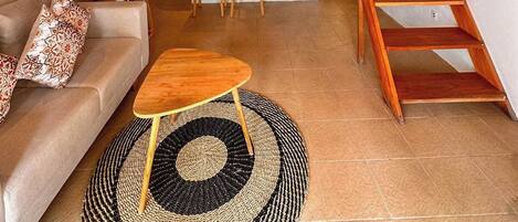 A cozy living area with a sofa, coffee table, and a woven rug. A wooden staircase leads to a kitchen with a bar area.