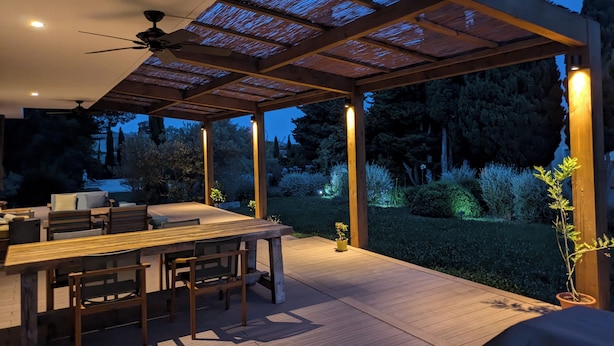 An outdoor patio with a wooden table and chairs, a ceiling fan, and a pergola with a translucent roof.