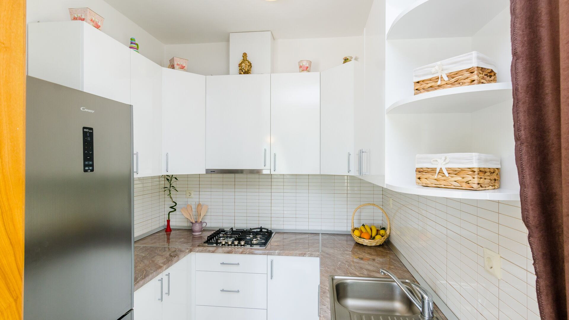 A modern kitchen with white cabinets, a built-in gas stove, a stainless steel refrigerator, and a basket of fruit on the counter.
