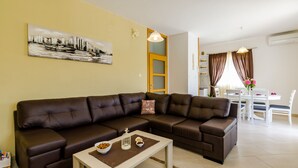 A modern living room with a brown leather sofa, a coffee table with decorative items, a dining area with a white table and chairs, and a cityscape painting on the wall.