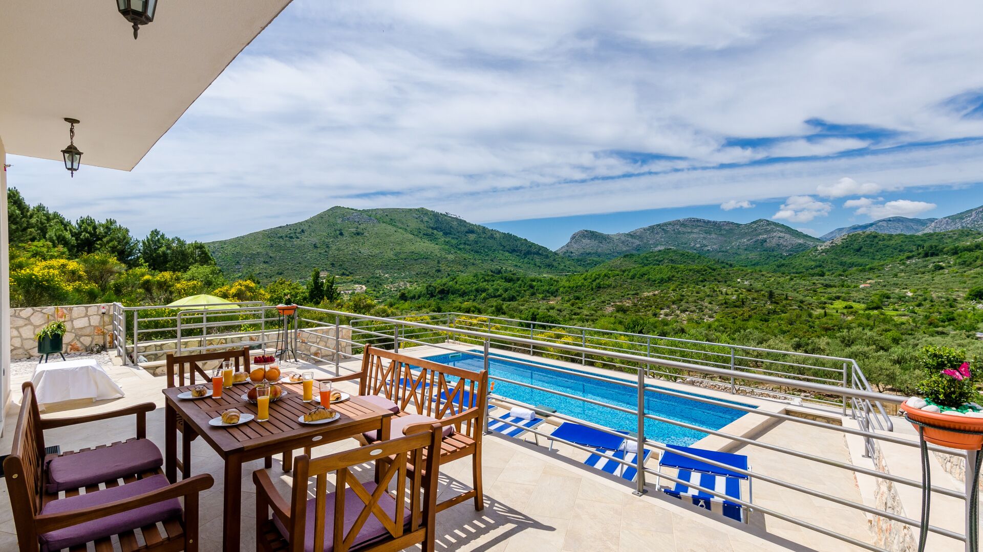A poolside area with a table and chairs, a pool, and a view of green hills.