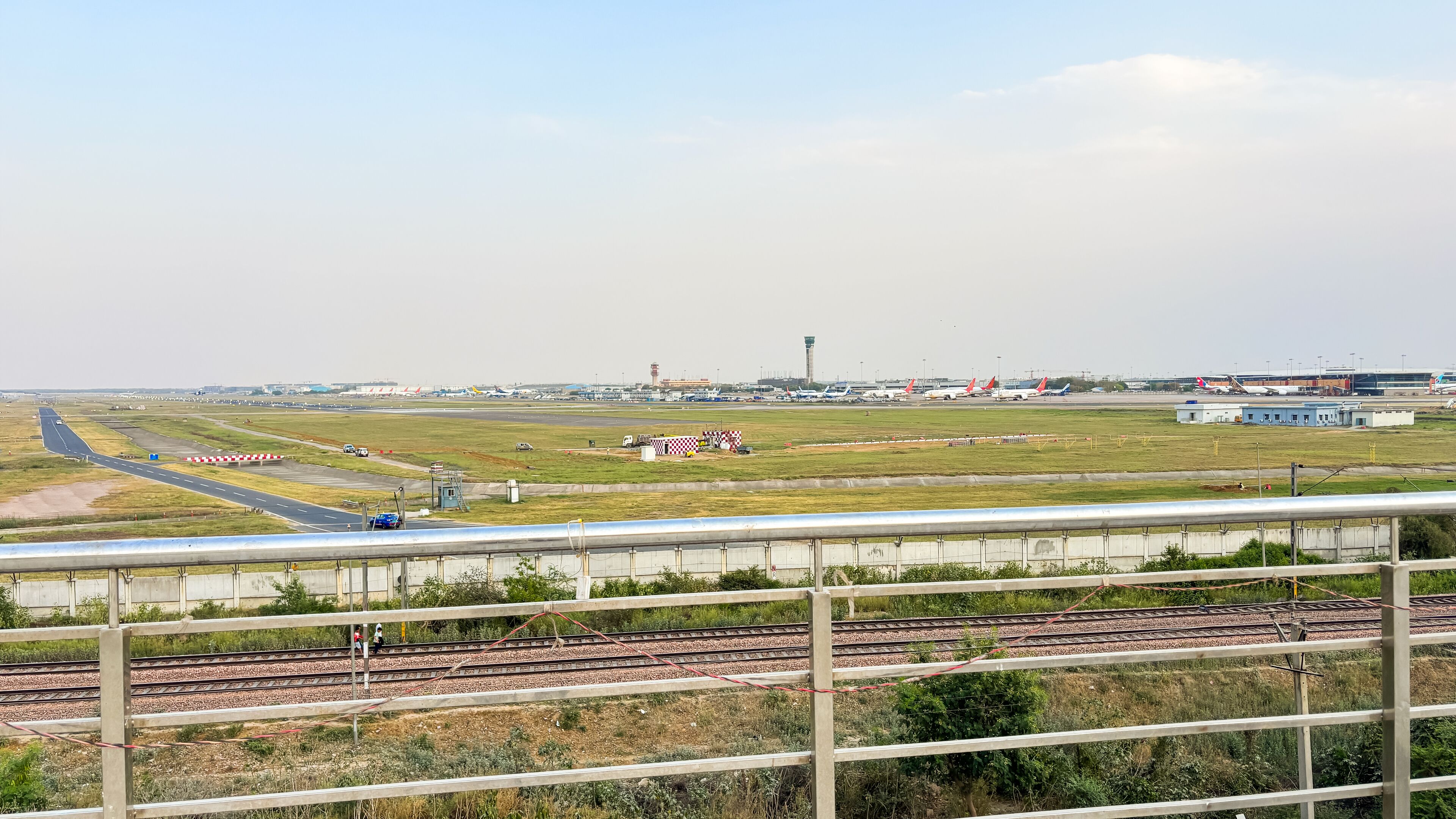 Une voie ferrée bordée d’une clôture, avec une vue lointaine d’un aéroport où se trouvent plusieurs avions et bâtiments.