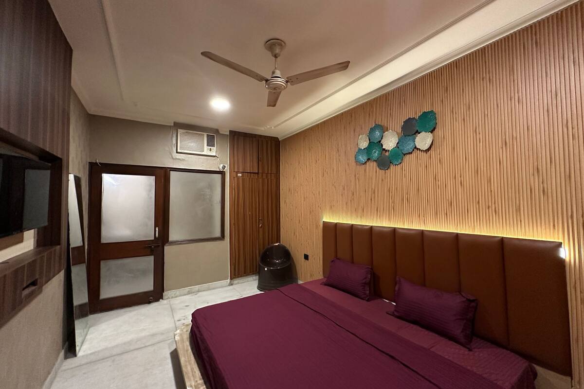 A bedroom with a bed, a ceiling fan, a wall-mounted air conditioner, and a decorative wall panel.