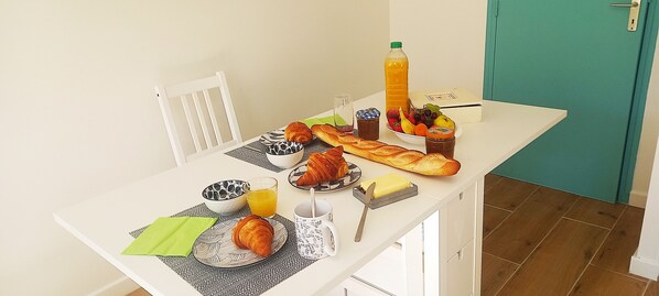 A breakfast table with croissants, a baguette, a bowl of fruit, a bottle of orange juice, and a glass of orange juice.