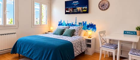 Ein Schlafzimmer mit Bett, Nachttischen, Schreibtisch, Stuhl, einer Uhr und einem an der Wand befestigten Fernseher, der Amazon Prime Video zeigt.