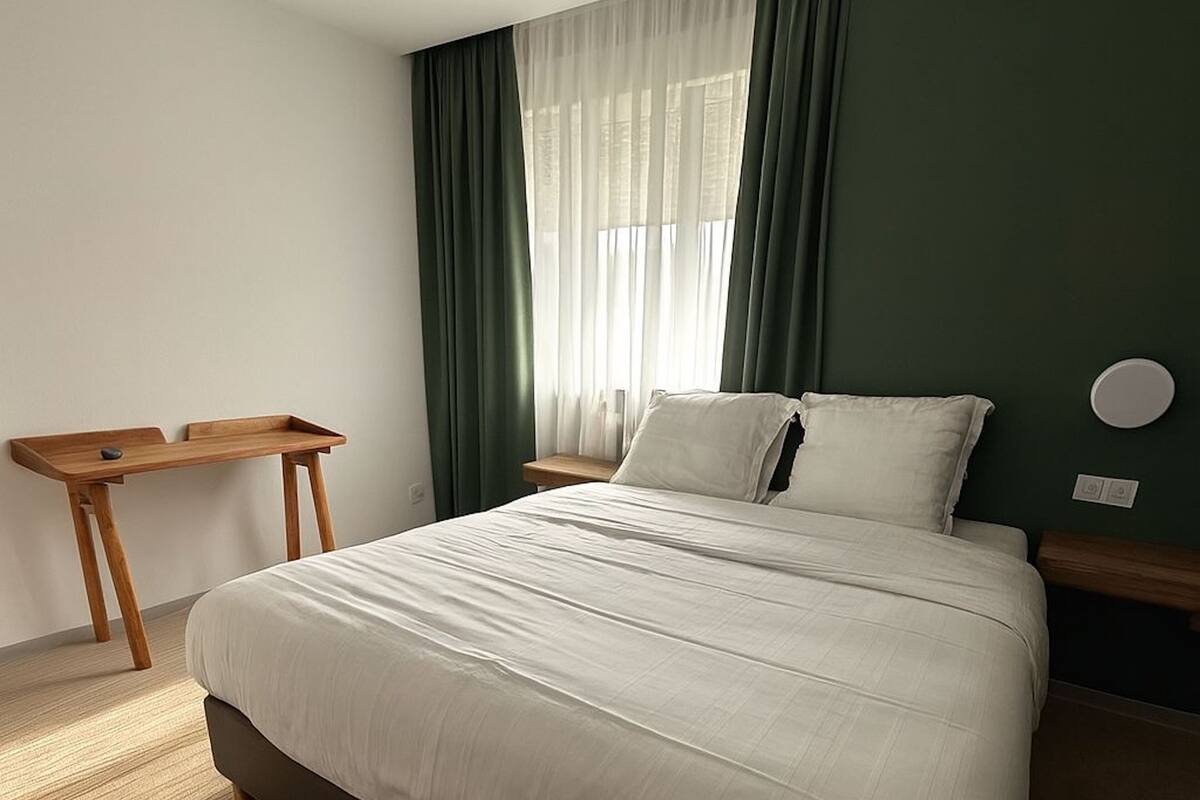 A neatly arranged bedroom with a bed, a wooden nightstand, a small table, and a window with sheer curtains.
