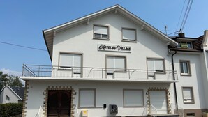 A two-story building with a sign reading 'HOTEL DU VILLAGE.' There are steps leading to the entrance, a small balcony on the upper floor, and a door with a window above it. - Hotel du Village (Village-Neuf)