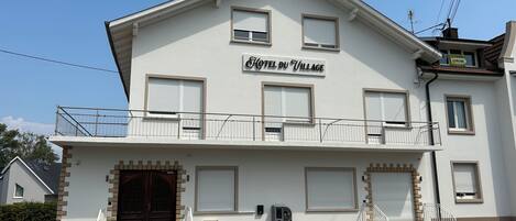 A two-story building with a sign reading 'HOTEL DU VILLAGE.' There are steps leading to the entrance, a small balcony on the upper floor, and a door with a window above it.