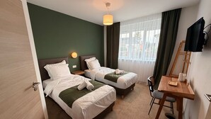 A hotel room with two beds, a desk with a chair, a television, and a window with curtains. - Hotel du Village (Village-Neuf)