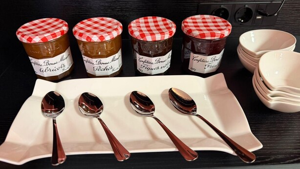 Four jars of jam with checkered lids on a black countertop, accompanied by four spoons and a stack of white bowls. - Hotel du Village (Village-Neuf)