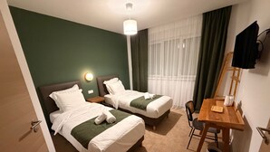 A hotel room with two beds, a desk, a chair, a lamp, and a window with curtains. - Hotel du Village (Village-Neuf)