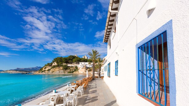 A seaside terrace with white tables and chairs, a blue window, and a view of the ocean and hills. - Casa Golondrina (Moraira)