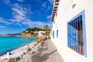 A seaside terrace with white tables and chairs, a blue window, and a view of the ocean and hills.