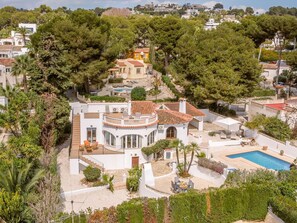 A Mediterranean-style villa with a pool, surrounded by lush greenery and a hilly backdrop.