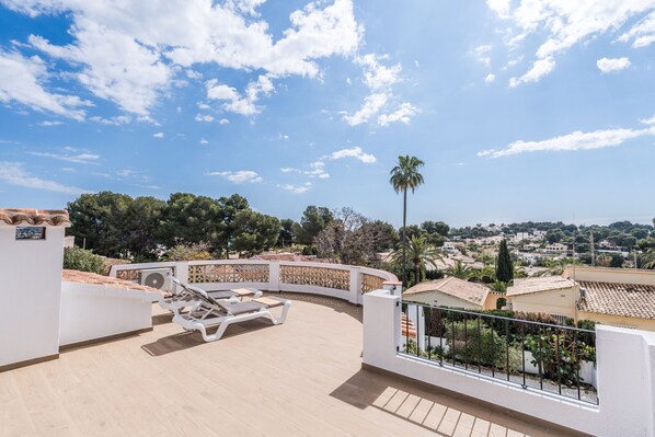 A rooftop terrace with lounge chairs, a palm tree, and a view of a town.