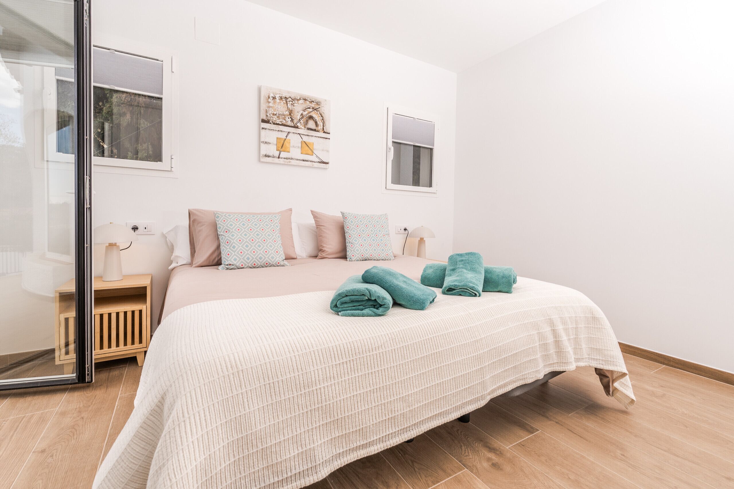 Een net opgemaakt bed met een witte deken en teal handdoeken, een nachtkastje van hout met een lamp, en twee ramen aan de muur bevestigd.