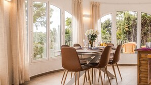 A dining area with a table set for four, surrounded by chairs, large windows, and a view of greenery. - Casa Golondrina (Moraira)