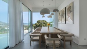 A modern dining room with a long table and multiple chairs, large windows with a view of the landscape, and contemporary artwork on the walls.