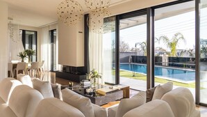 A spacious living room with a large sofa set, a coffee table, and a fireplace. There are large windows offering a view of a swimming pool and outdoor area.