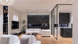 A modern living room with a white sofa, a coffee table, and a TV. There is a stone wall and a kitchen area visible through a glass door.