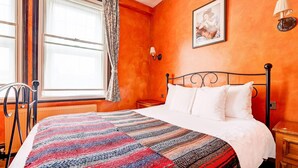 A bedroom with a bed, a window with curtains, a radiator, a bedside table, and a wall-mounted lamp.