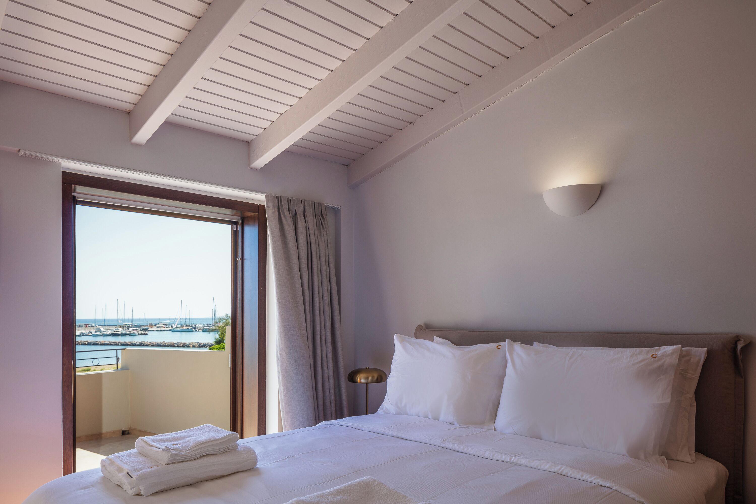 Habitación de hotel con cama, mesita de noche, lámpara, vista al puerto deportivo y balcón.