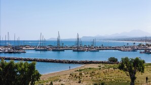 A marina with boats docked, a rocky breakwater, and a clear view of the sea and distant mountains.