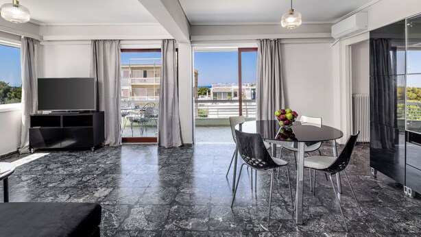 A modern living room with a flat-screen TV, a dining table with chairs, and a balcony with a view of the city. - Seaside Secret Sea View Two Bedroom Apartment by BluPine (Athens)