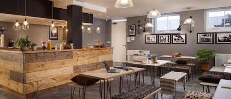 Un intérieur moderne avec un comptoir central en bois, de nombreuses tables et chaises, des luminaires suspendus et des tableaux encadrés aux murs.