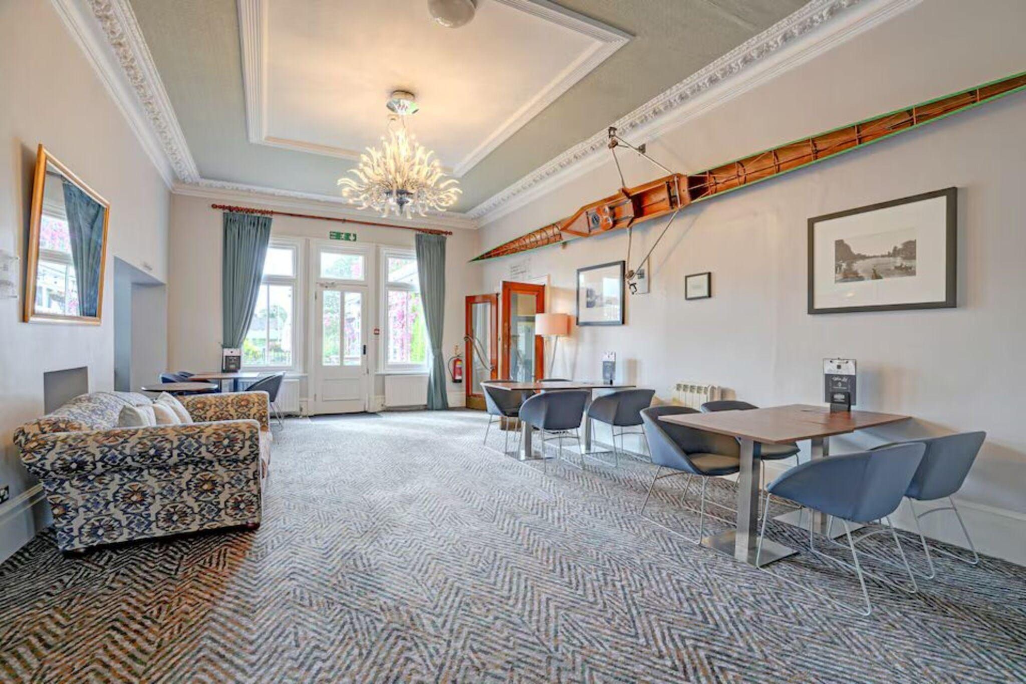 A hotel room with a patterned carpet, a sofa, a dining table with chairs, and a chandelier.