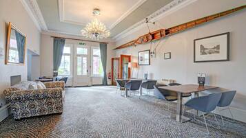 A hotel room with a patterned carpet, a sofa, a dining table with chairs, and a chandelier.