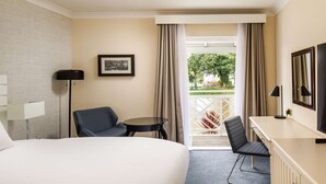 Ein Hotelzimmer mit Bett, zwei Stühlen, einem Schreibtisch, einem Fernseher und einem Fenster mit Vorhängen.